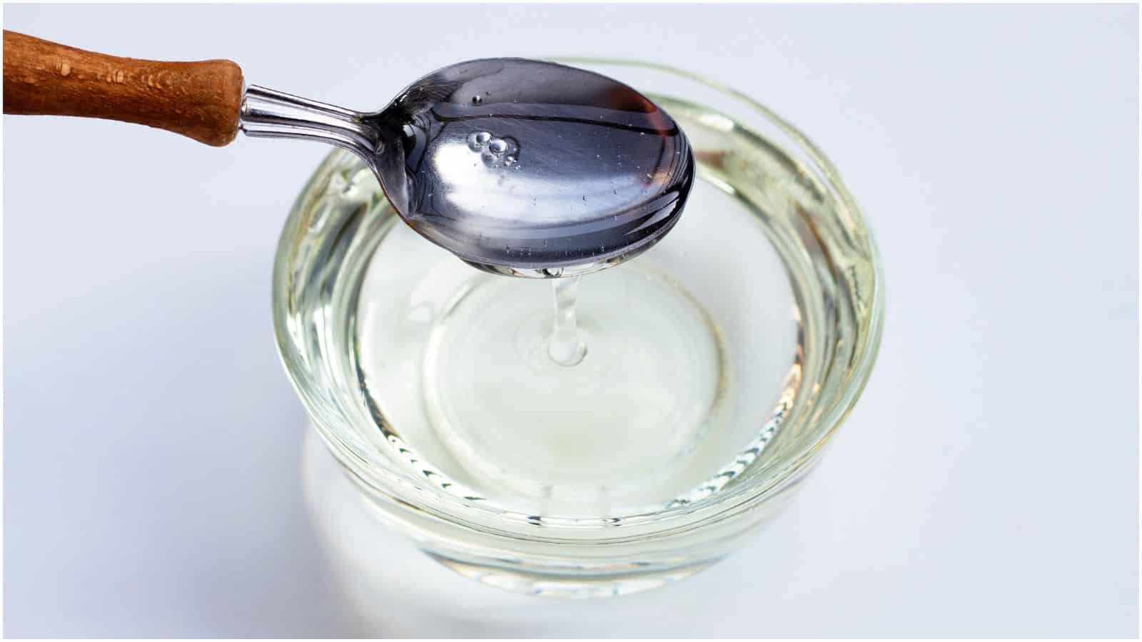A spoon held over a glass container filled with clear liquid, some of it dripping back in. It's everything you need to know about simple syrup captured in a minimalist shot against a plain white background.