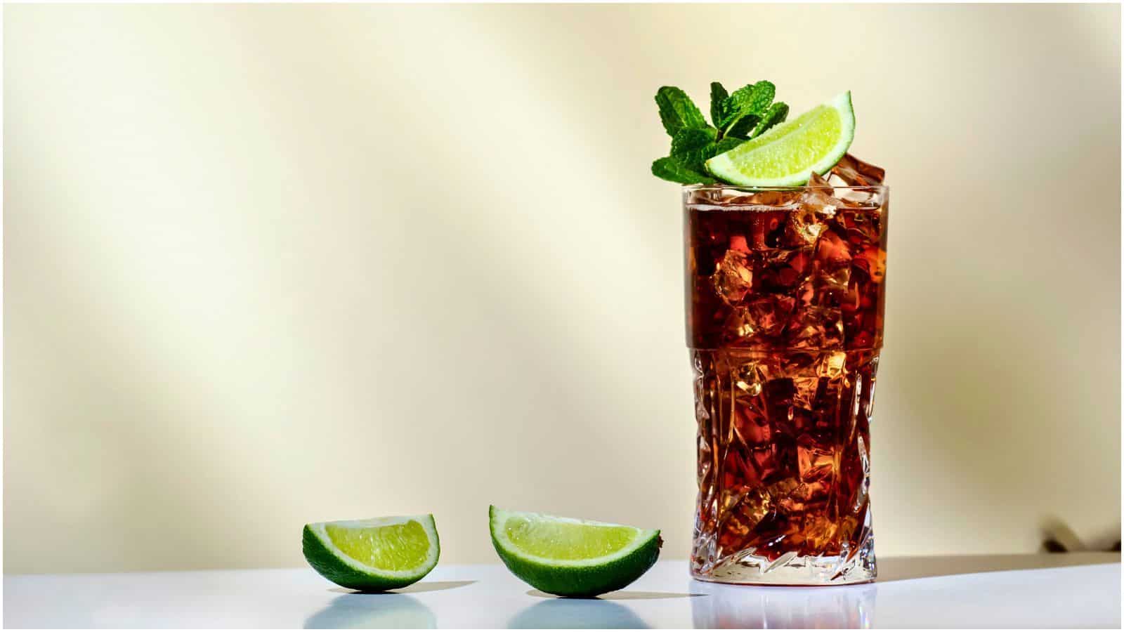 A tall glass of iced beverage, possibly an alcoholic drink that is bad for your teeth, garnished with a lime wedge and mint leaves. The glass contains ice cubes and a dark liquid. Two lime wedges are placed on the white surface in front of the glass. The background is blurred.