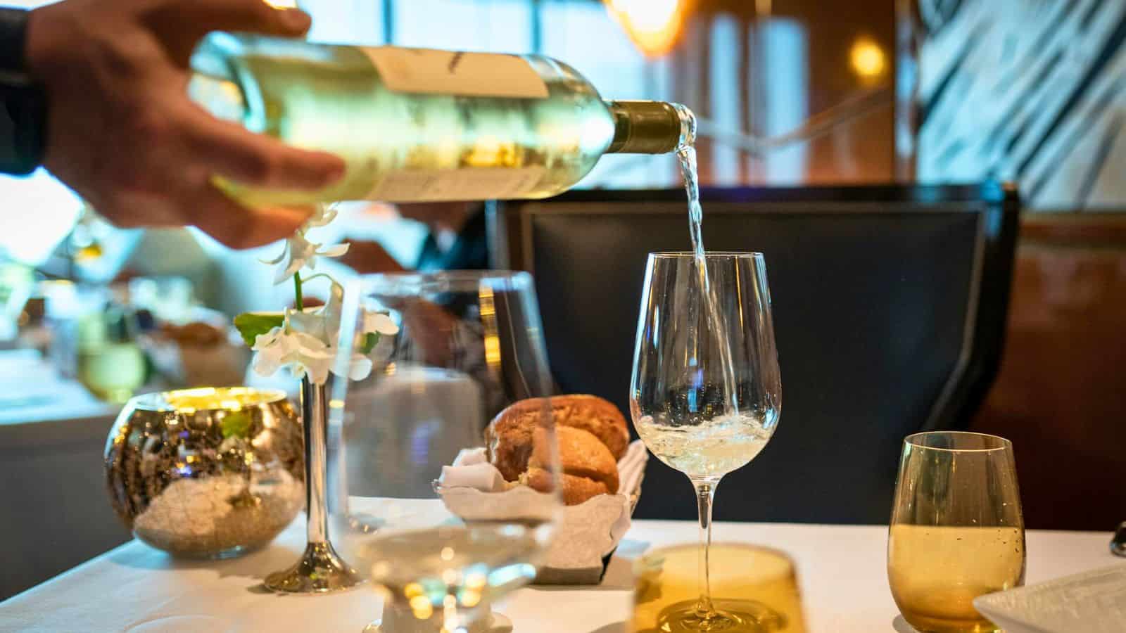 A person is pouring white wine, one of the alcoholic drinks that are bad for your teeth, from a bottle into a wine glass at a restaurant table. The table is set with other wine glasses, a bowl of bread rolls, and various decorative items. The background shows a stylish dining area.