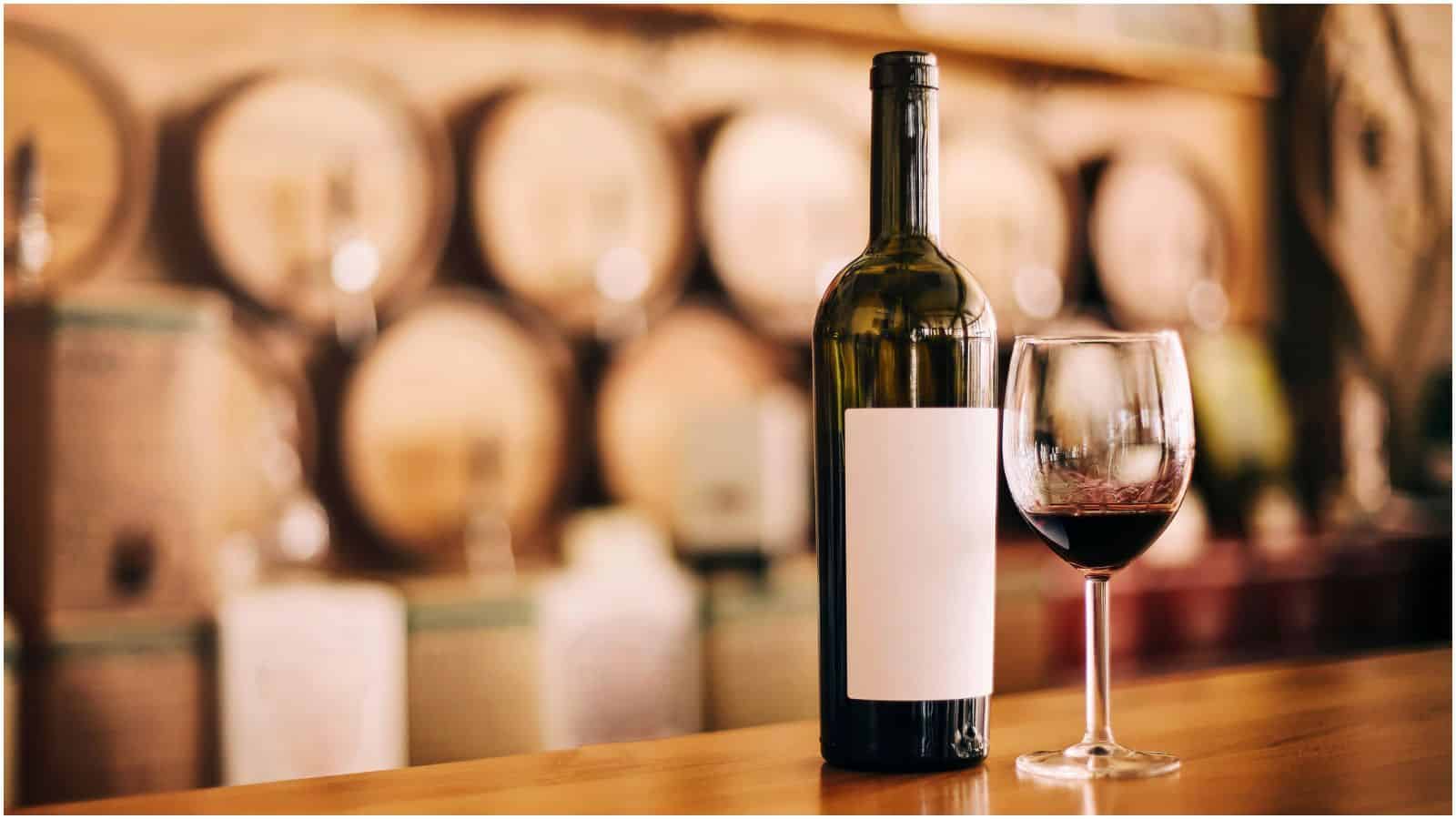 Bottle of wine and filled wine glass with wine barrels in the background