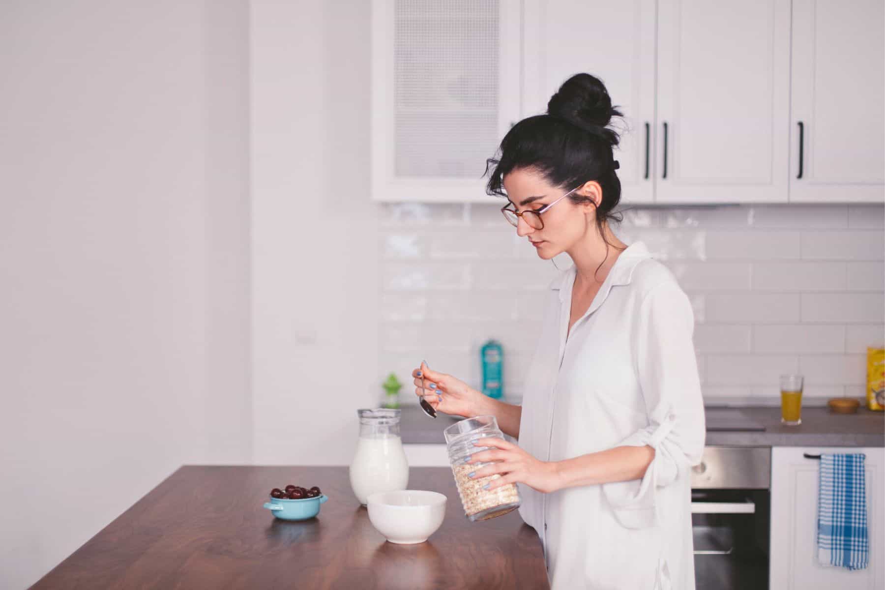 A woman with glasses and a bun is standing in a kitchen, pouring cereal into a white bowl. There is a blue bowl with fruit, a glass jug with milk, and a glass of orange juice on the wooden counter. White cabinets and a tiled backsplash are in the background.