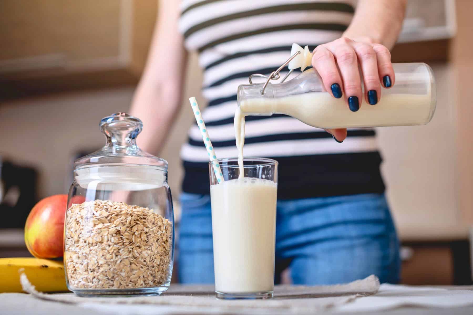 A person is pouring milk from a glass bottle into a glass with straws on a kitchen counter. Next to the glass is a jar filled with oats and an apple. The person is dressed in a striped shirt and jeans.