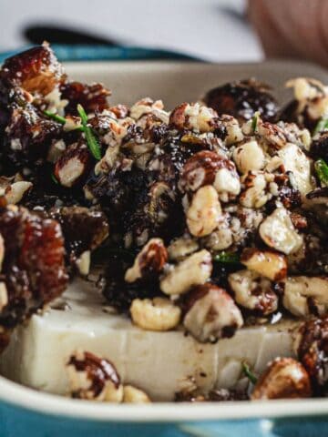 A close-up of a dish featuring a block of feta cheese topped with a mix of chopped nuts and herbs. Two hands with light pink nails are holding the dish, which is placed in a light blue rectangular baking tray.