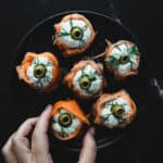 A black plate with six smoked salmon rolls topped with white cheese, green olives, and dill. A hand is holding one of the salmon rolls near the bottom left corner of the plate. The background is black.
