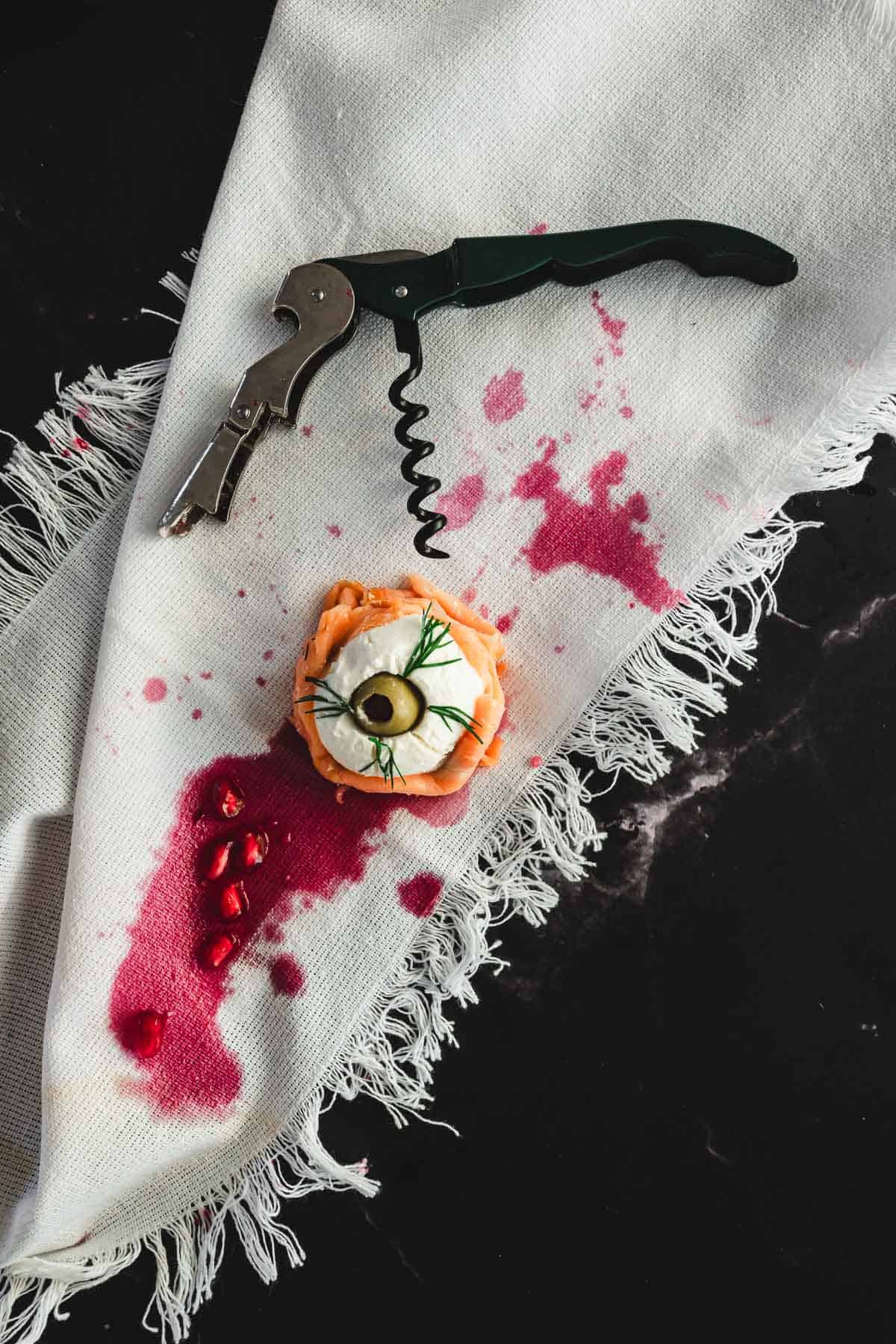 A corkscrew rests on a white fringed cloth stained with red liquid. Beside it, a Halloween-themed canape sits—a small pastry topped with cream, smoked salmon, dill, an olive, and red berry seeds—creating a mysterious scene against the dark surface.