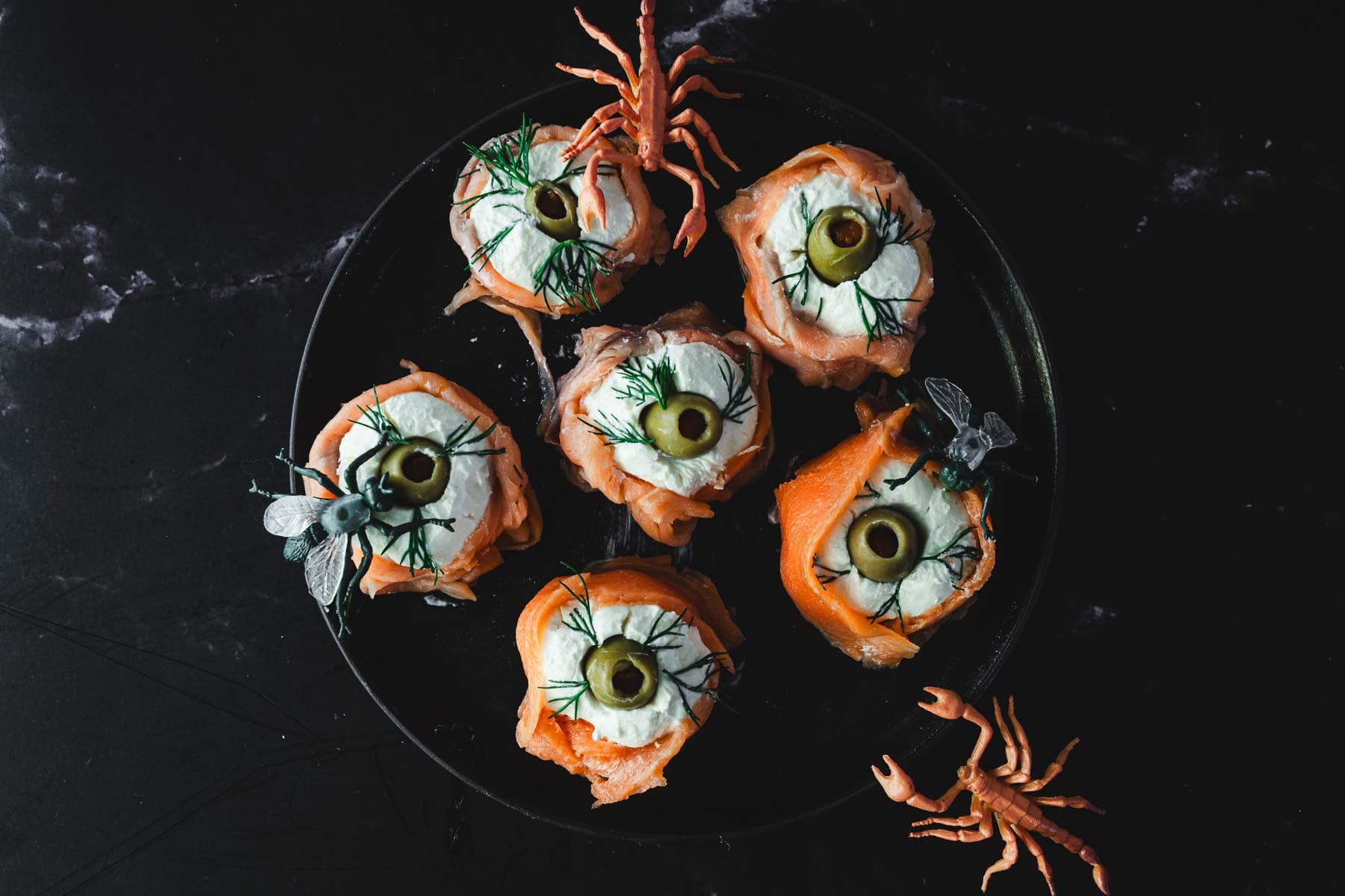 A black plate holds six Halloween smoked salmon canapes, each featuring cream cheese wrapped with salmon and topped with green olives and dill. Decorative plastic insects crawl around the plate on a dark marble surface, adding a spooky charm.
