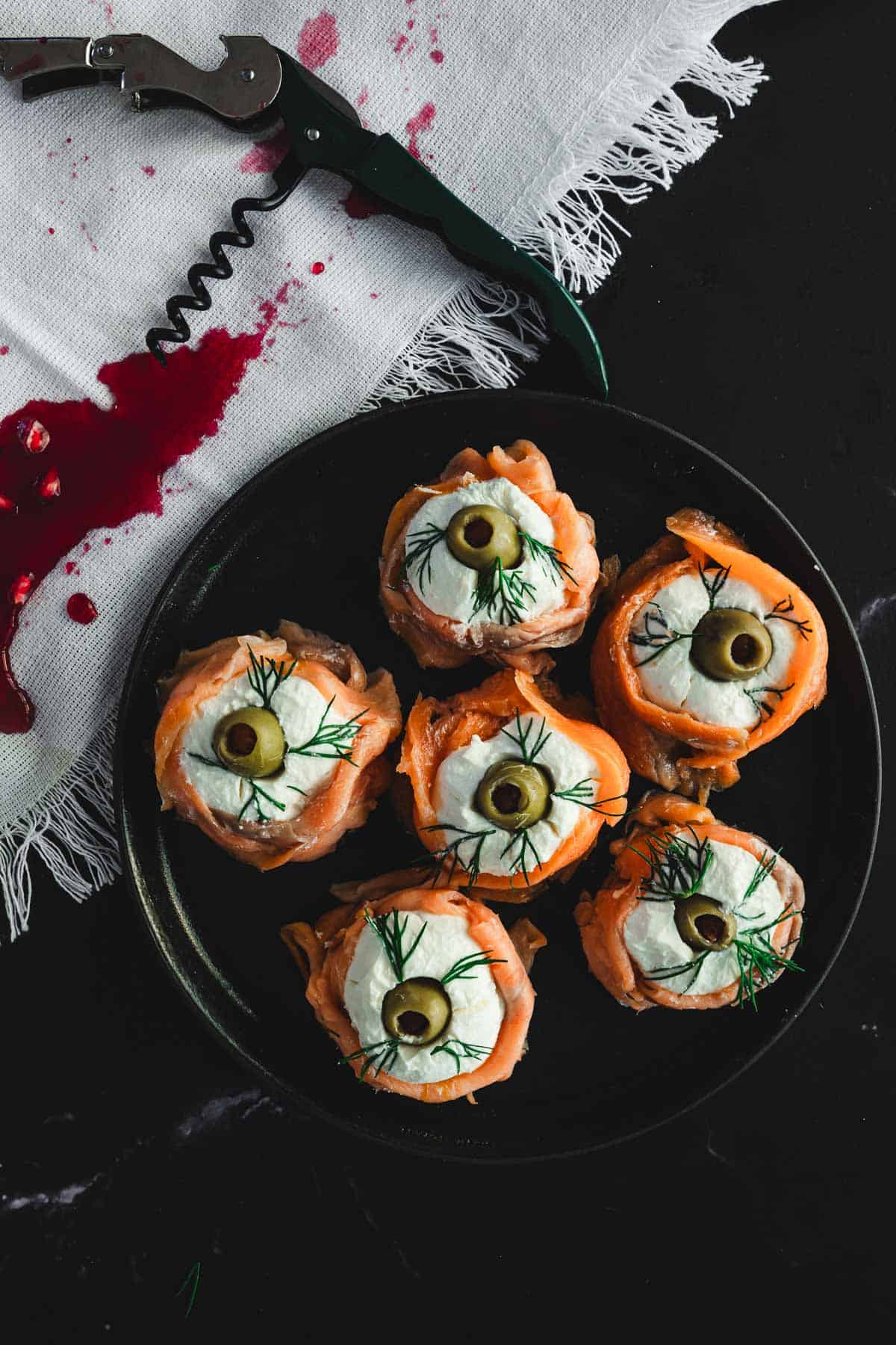 A plate with six Halloween smoked salmon canapés resembling eyes, crafted from smoked salmon, cream cheese, a green olive slice, and dill. A corkscrew and a cloth with a red stain rest beside it on the dark surface.