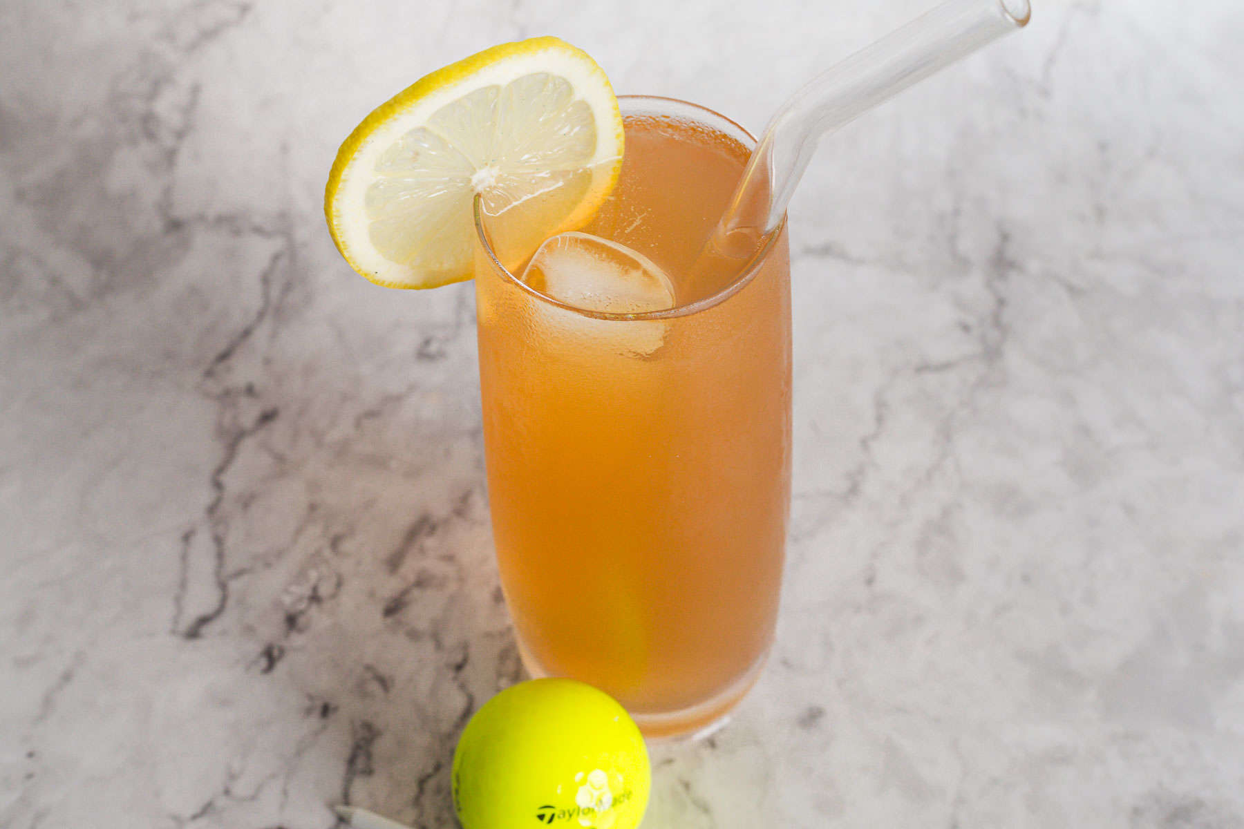 A tall glass of iced peach-colored drink with a lemon slice garnish and a glass straw on a marbled surface. A yellow plastic golf ball lies nearby.