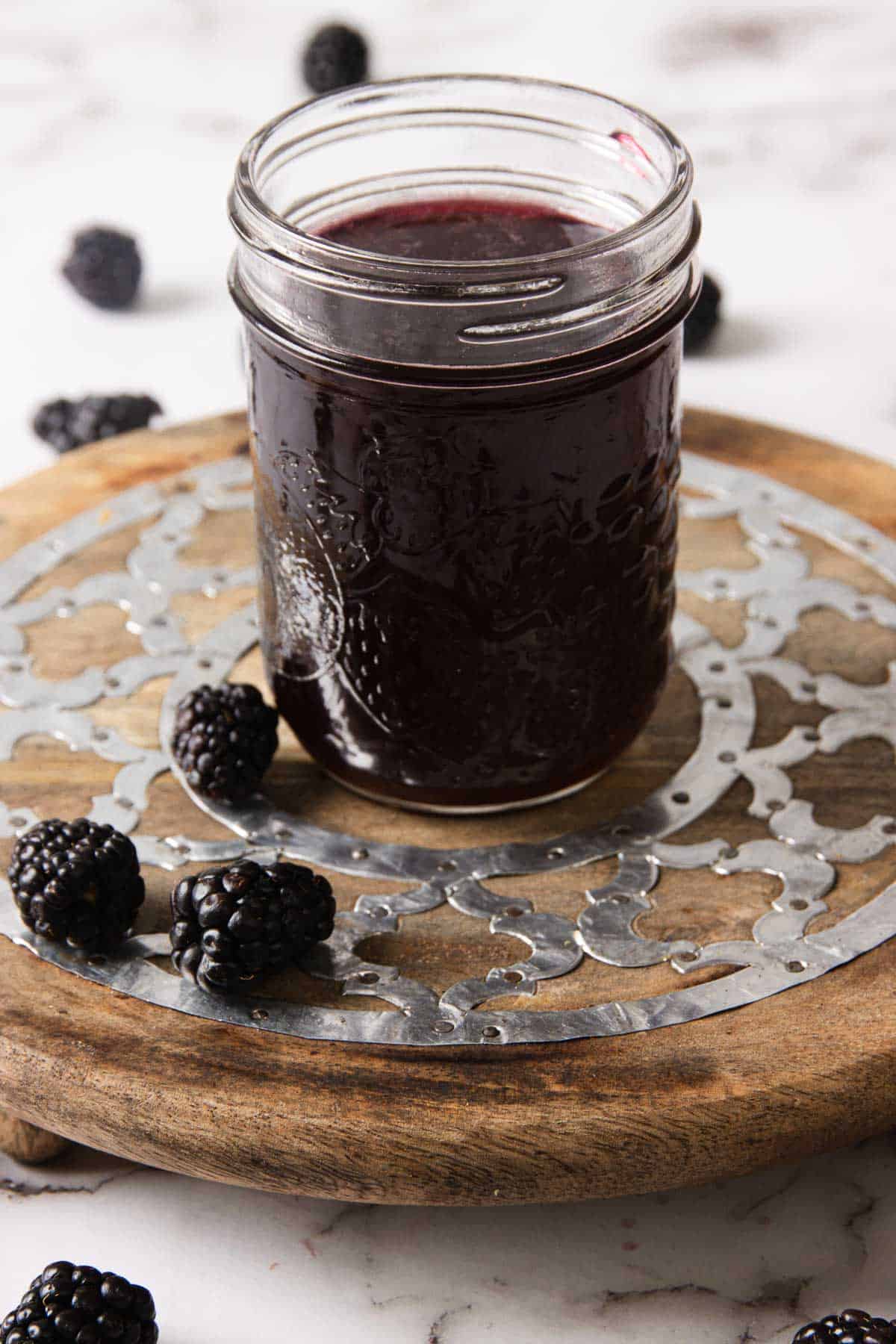 A glass jar filled with dark purple blackberry jam is placed on a round wooden coaster with a metal decorative insert. Several blackberries are scattered around the jar on the marble surface.