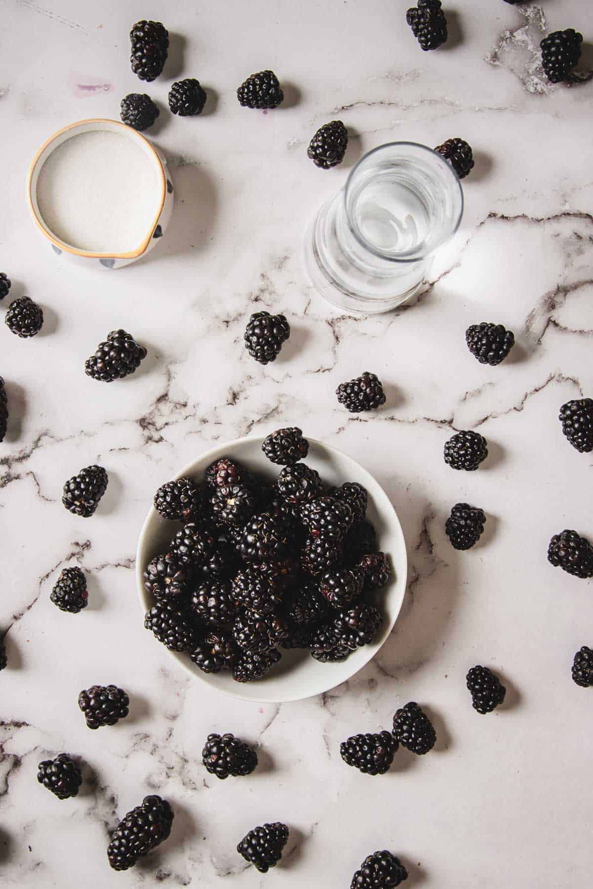 A white bowl filled with blackberries is placed on a marble surface. Scattered blackberries surround the bowl along with a small bowl of sugar and a glass of water.