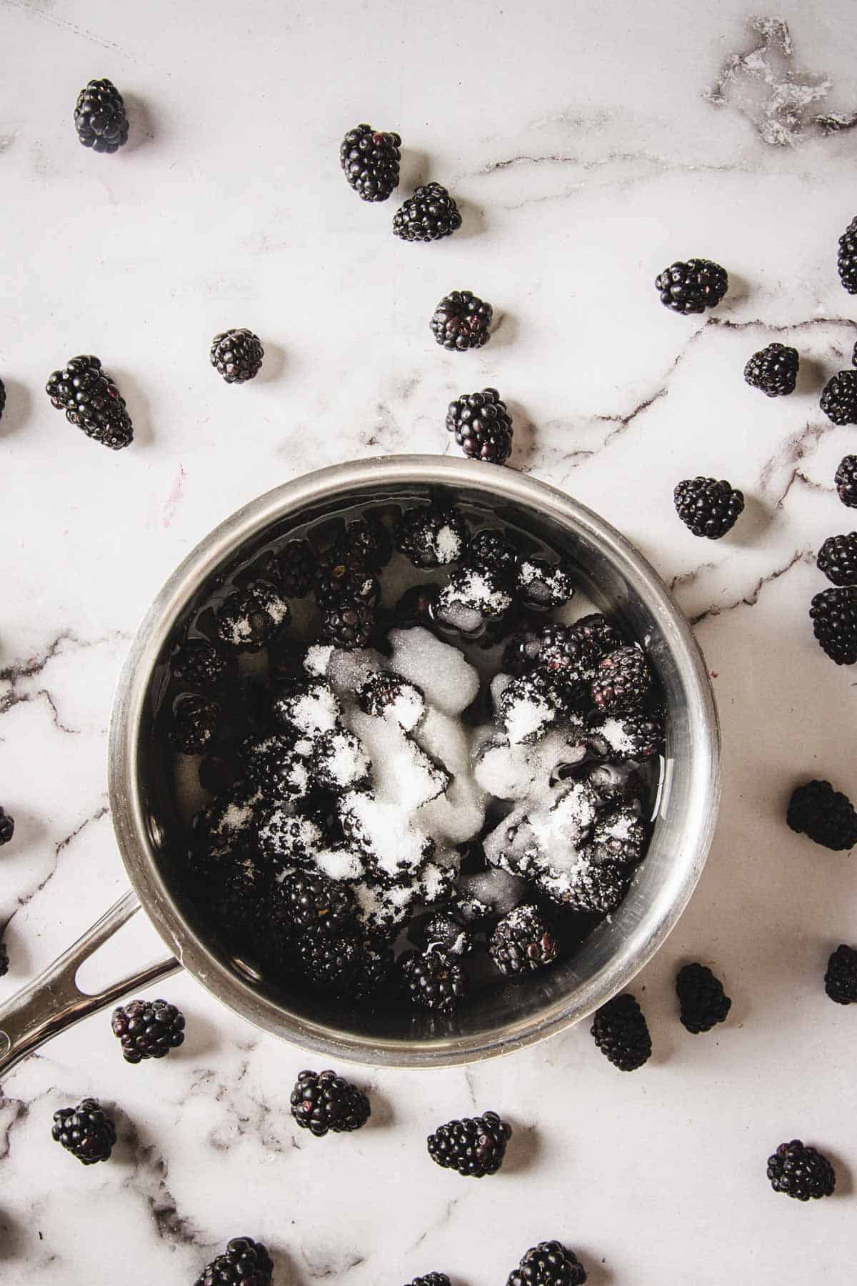 A stainless steel pot filled with blackberries that are partially covered in sugar sits on a marble countertop. Several blackberries are scattered around the pot on the countertop.
