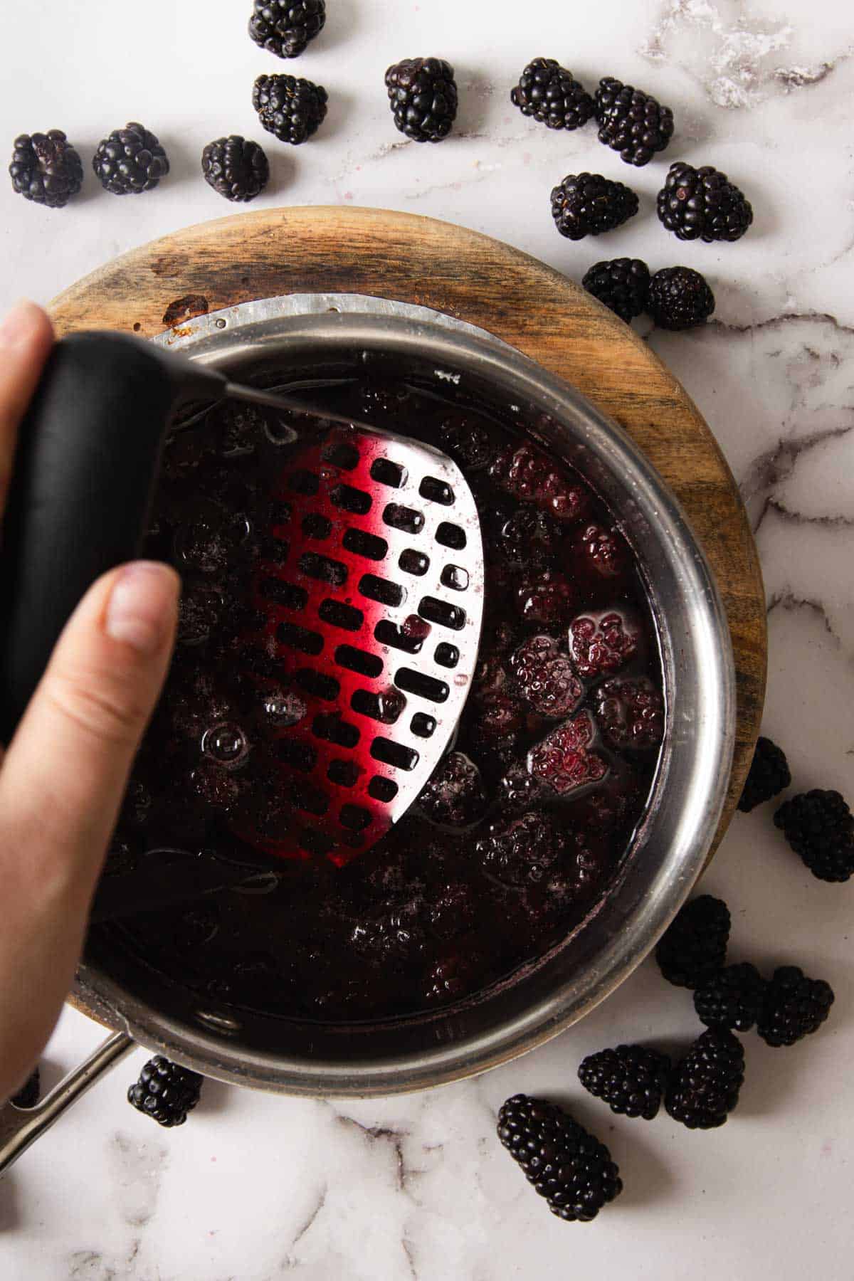 A hand using a potato masher to crush blackberries in a stainless steel pot on a round wooden board. Fresh blackberries are scattered on a white marble countertop around the pot.