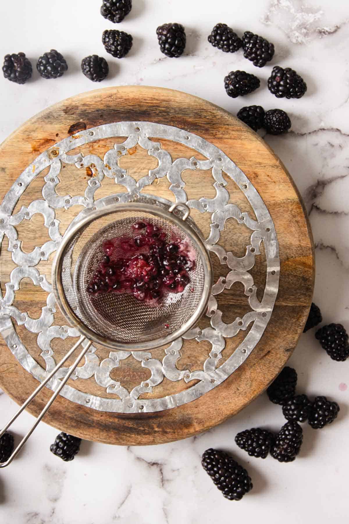 A round metal strainer filled with crushed blackberries sits on a mandala-like wooden board atop a marble surface. Whole blackberries are scattered around the strainer and on the marble surface.
