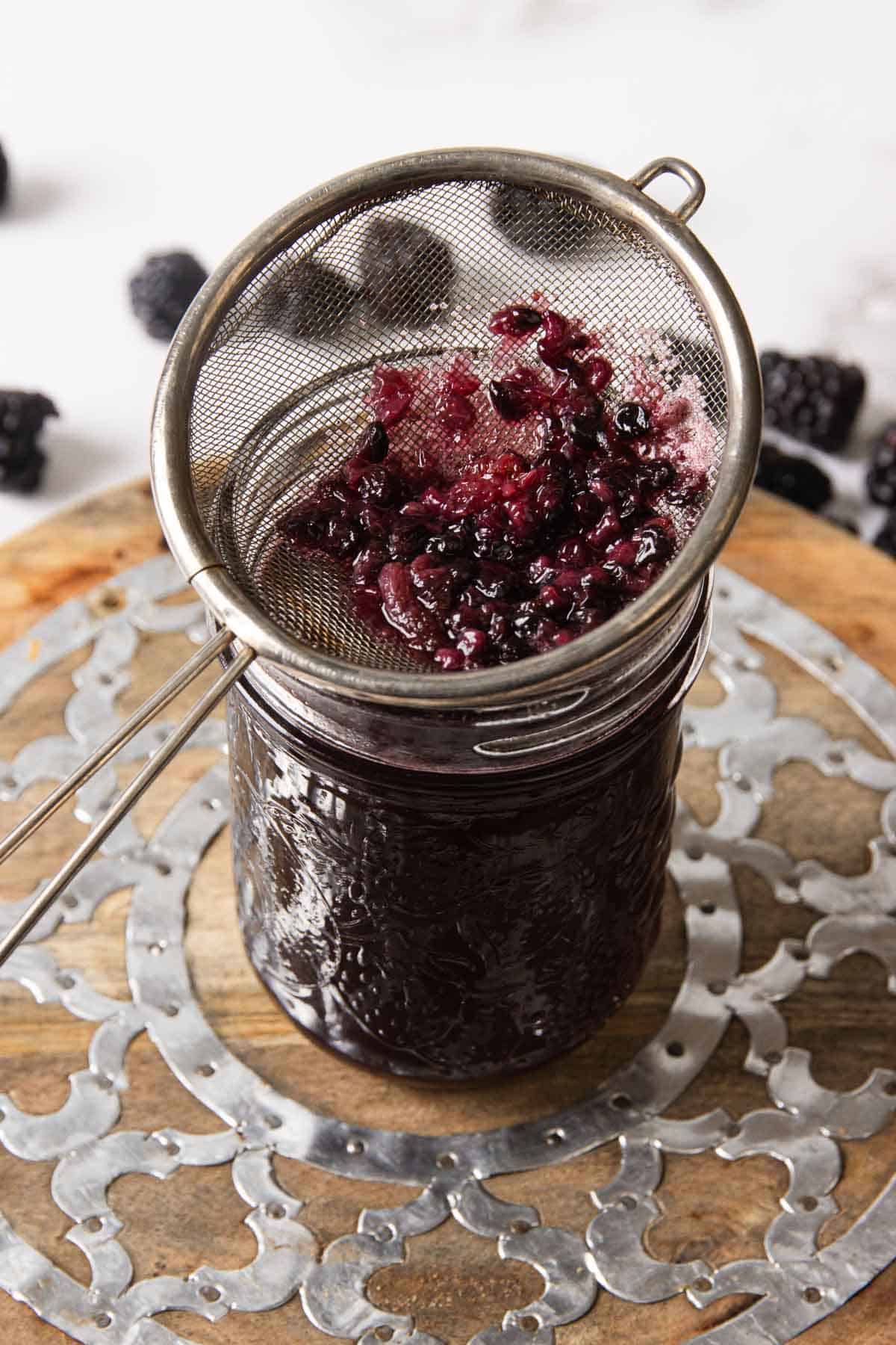 A glass jar filled with dark liquid and a fine mesh strainer placed on top, containing crushed blackberries. The jar is on a decorative wooden and metal tray, with more blackberries scattered in the background.