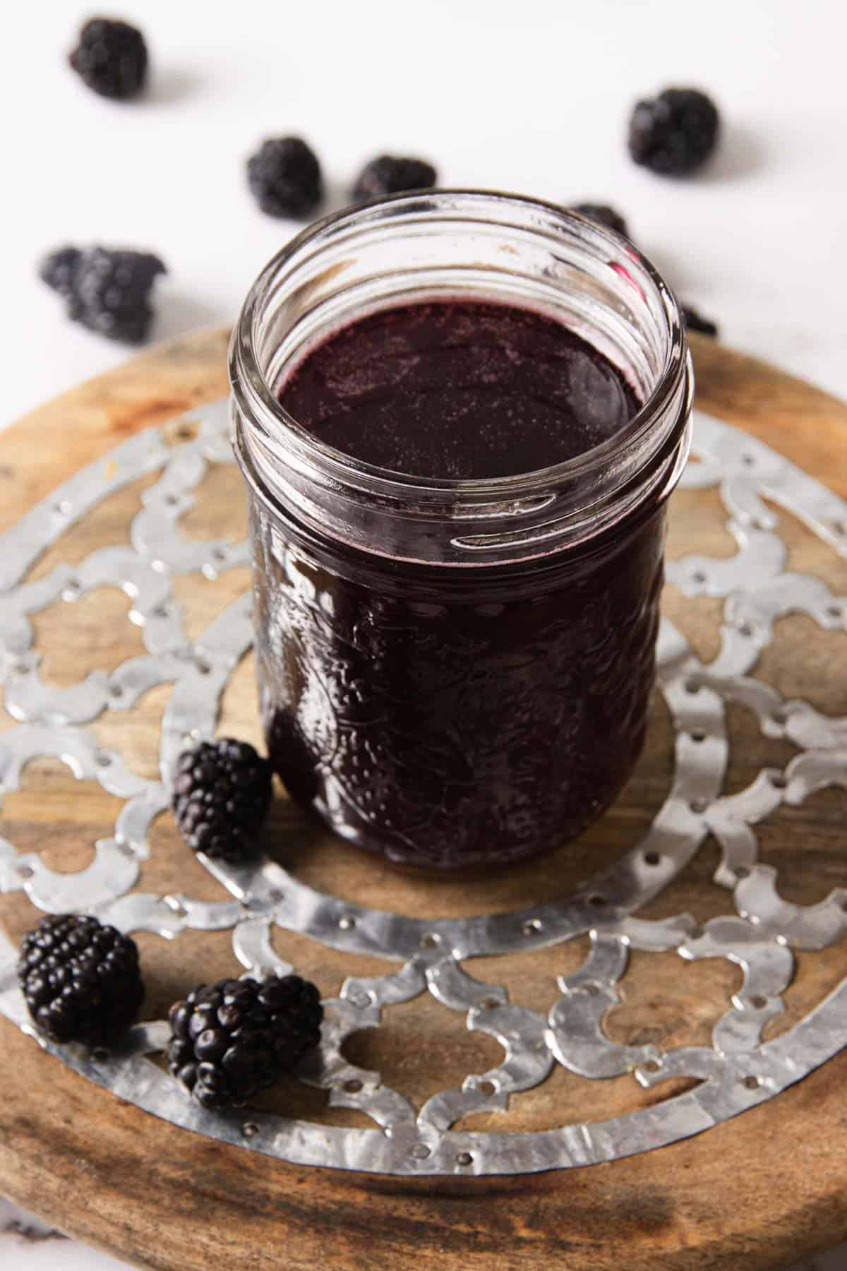 A glass jar filled with blackberry jam is placed on a round wooden surface with a decorative metal overlay. Fresh blackberries are scattered around the jar. The background is blurred and white.