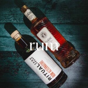 Two bottles of rum are displayed on a dark wooden surface. The bottle at the top is labeled ÉLITE RUM with a pattern on the glass. The bottle at the bottom is labeled RITUAL ZERO PROOF, featuring a minimalist design with primarily white and red labels. The word "rum" is centered in white text between the bottles.