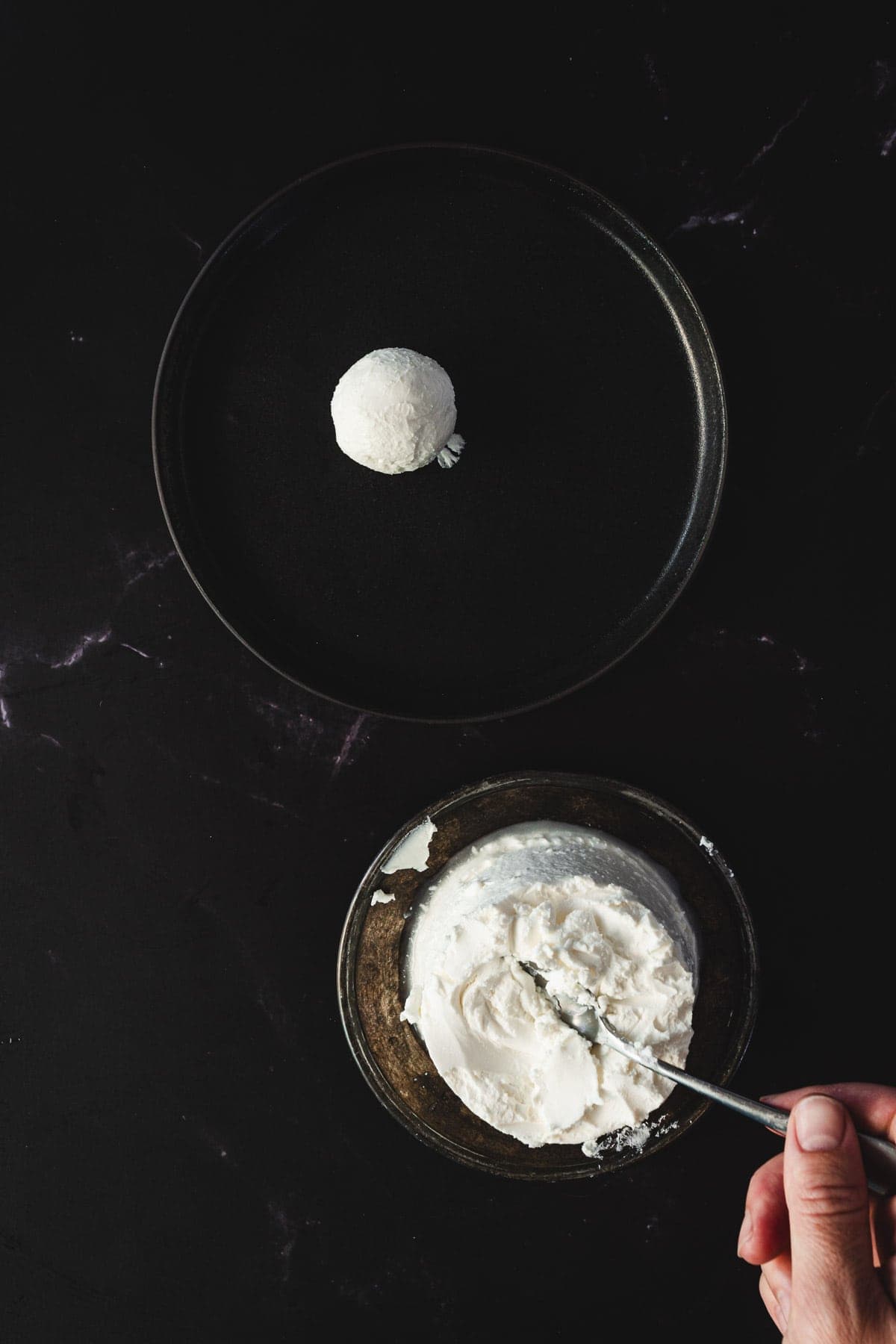 A hand holds a spoon over a bowl of soft white cheese on a dark surface, ready to craft Halloween smoked salmon canapés. A small scoop of cheese rests on an adjacent black plate. The overhead view captures the minimalist scene perfectly.