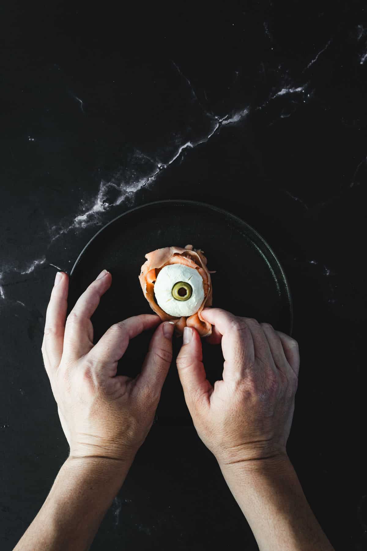 Two hands are placing Halloween smoked salmon canapés on a dark plate against a dark marble surface. Each appetizer showcases a slice of salmon wrapped around a festive filling, topped with cucumber and an olive.