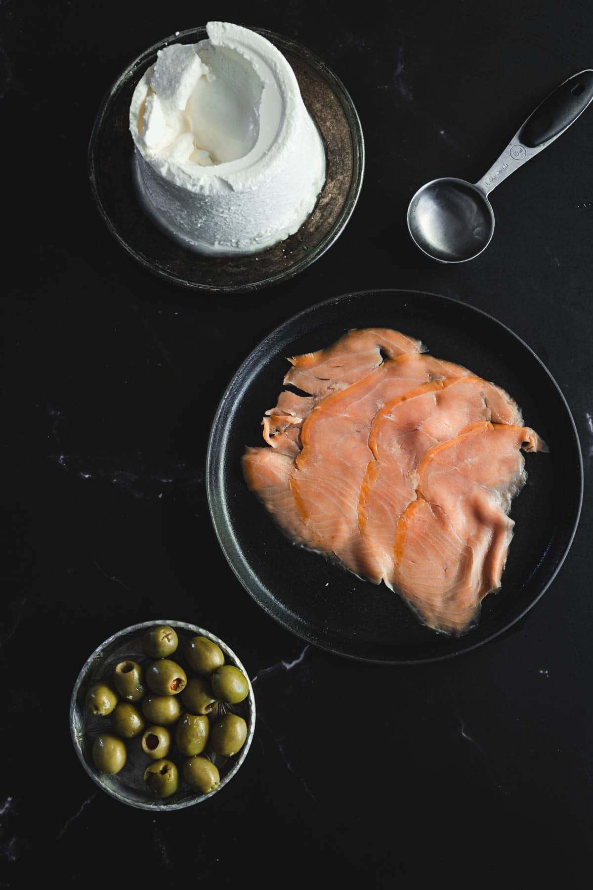 A dark surface sets the stage for a Halloween feast, featuring a black plate of smoked salmon slices ready to become canapés. A black bowl with goat cheese and a small bowl of green olives complete the eerie tableau, while a metal measuring cup waits nearby.