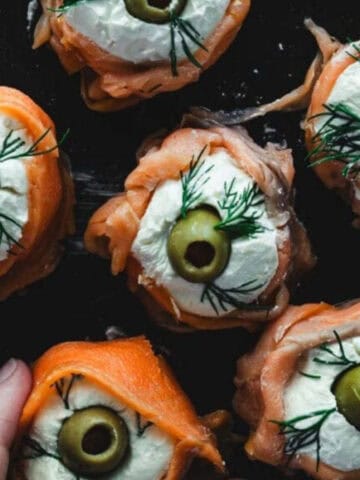 A close-up of several smoked salmon appetizers arranged on a dark plate. Each appetizer consists of smoked salmon, a dollop of cream cheese, a green olive slice, and a small sprig of dill. A hand is holding one of the appetizers.