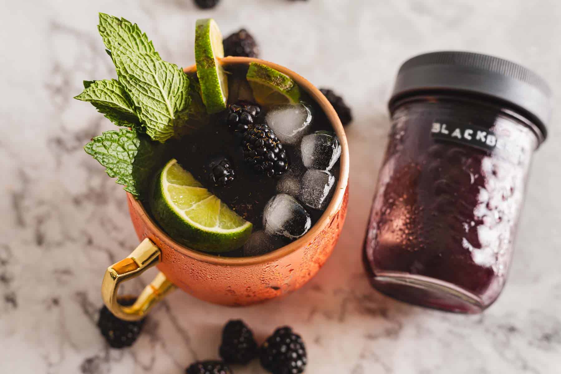 An overhead view of a copper mug filled with a vibrant blackberry Moscow Mule mocktail, featuring ice, blackberries, lime slices, and mint leaves. A clear jar with a black lid labeled "blackberry" sits beside the mug, while loose blackberries are artfully scattered around.