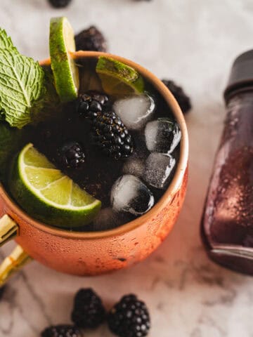 A copper mug filled with a dark blackberry Moscow mule mocktail, garnished with blackberries, lime slices, and mint leaves, sits on a marble surface. Ice cubes float in the drink. Beside the mug is a small jar labeled "Blackberry.