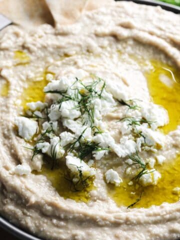 A close-up of a bowl of simple white bean hummus garnished with crumbled feta cheese, olive oil, and dill. Part of the bowl contains pita chips, and a small dish with salt and pepper is visible in the background. The bowl is placed on a wooden surface.