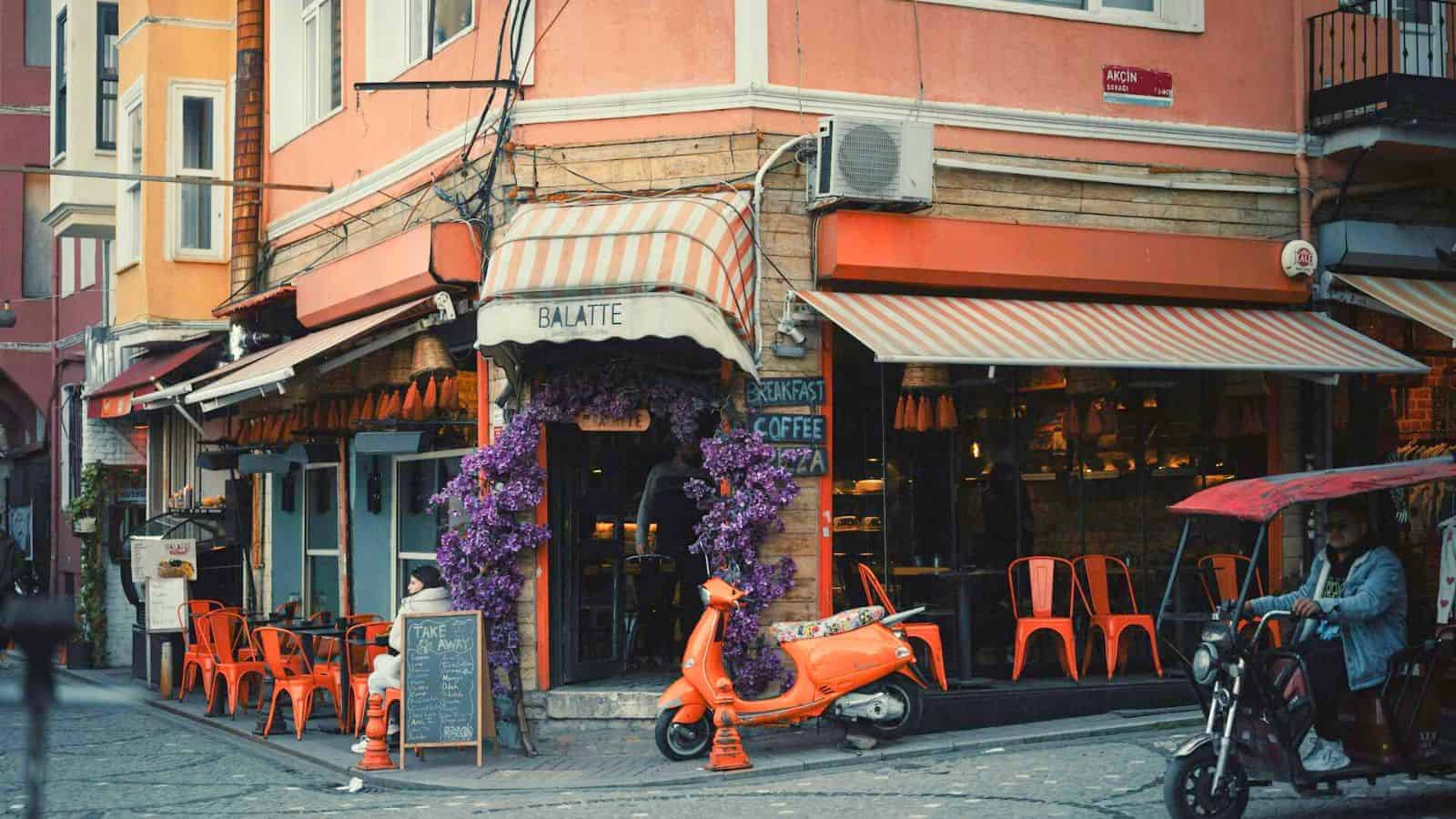 A street scene featuring a corner caf&eacute; with orange chairs and striped awnings. A vintage orange scooter is parked outside, adorned with purple flowers. A sign displaying "Breakfast & Coffee" stands beside the entrance.