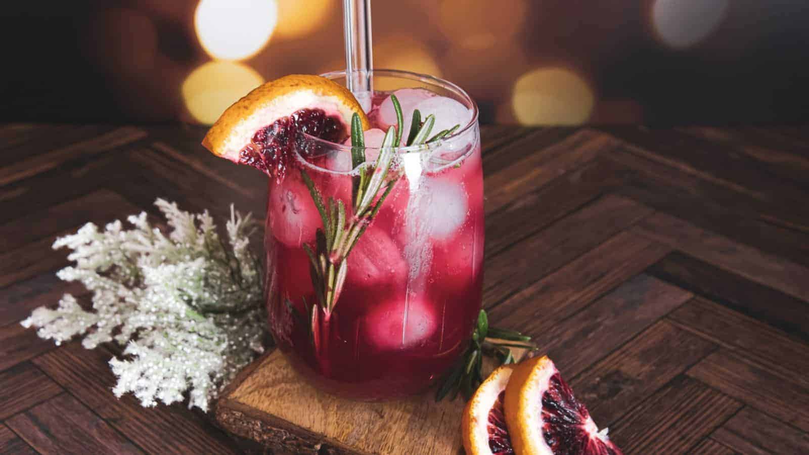 A glass of red cocktail with ice, garnished with rosemary and a slice of blood orange, sits on a wooden surface. A sprig of white decorative foliage and additional blood orange slices are nearby.