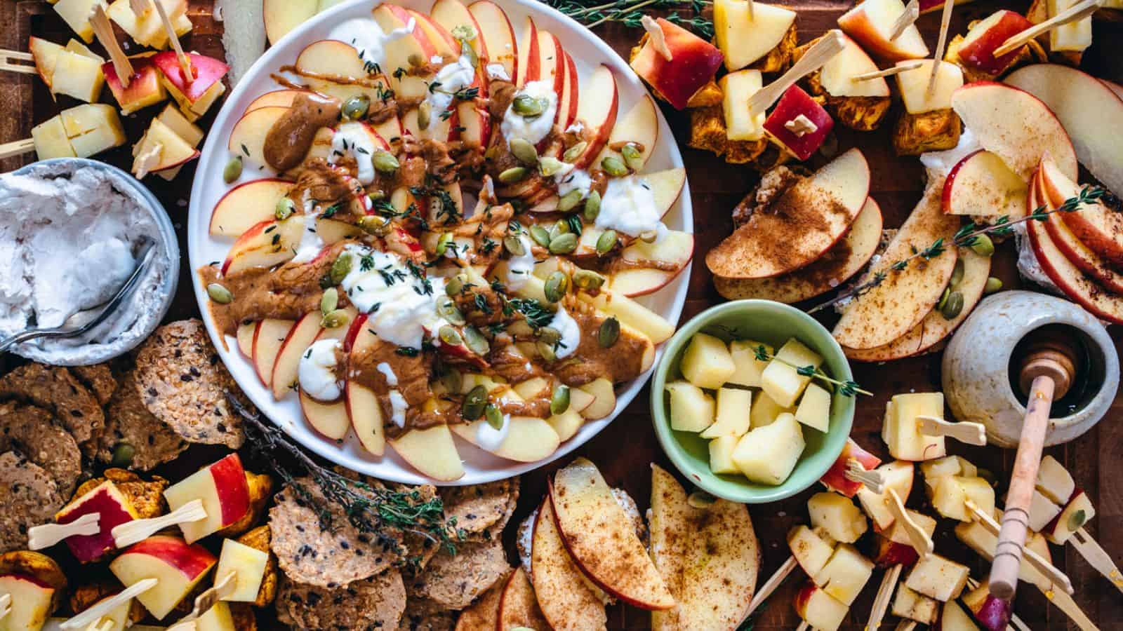 A platter of sliced apples topped with yogurt, seeds, and a drizzle of nut butter, surrounded by various snacks including cheese cubes, apple and cheese skewers, seed crackers, and a small bowl of pineapple chunks on a wooden table.