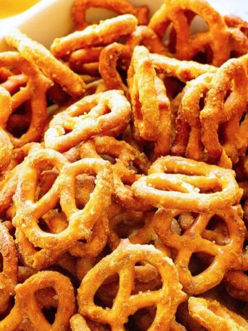 A white bowl filled with crispy, golden brown pretzels. The pretzels have a classic knotted shape and a lightly salted appearance. They are placed on a surface with a yellow cloth partially visible in the background.