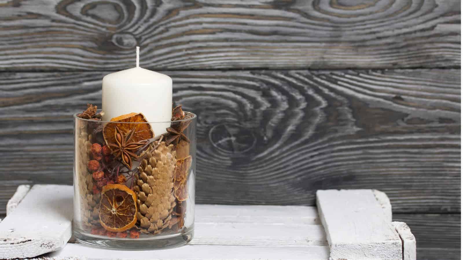 A white candle sits atop a glass filled with dried fruits, pinecones, and spices. The glass is placed on a white wooden surface. The background features gray, wooden planks with a prominent grain pattern.