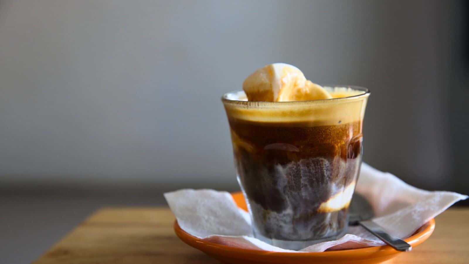 A glass of affogato on a small orange saucer with a spoon. The affogato consists of espresso poured over vanilla ice cream, creating a contrast of dark coffee and creamy white ice cream. It rests on a wooden surface with a blurred gray background.