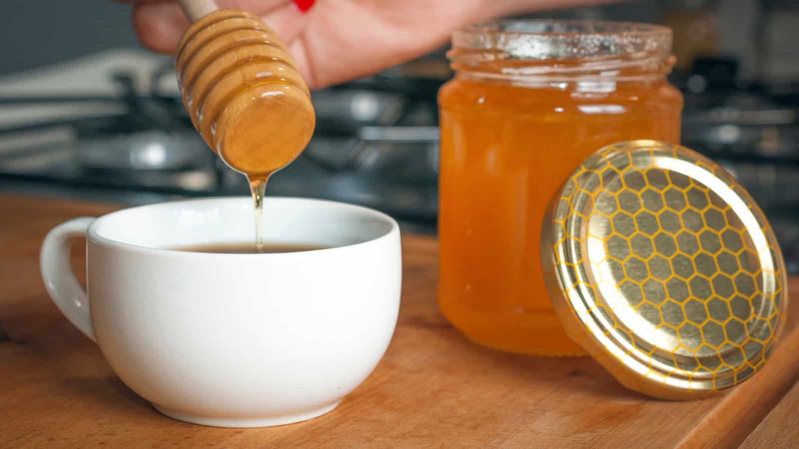 A hand drizzles honey from a wooden dipper into a white cup, much like the subtle sweetness found in underrated Starbucks flavors. Beside the cup sits an open jar of honey with a gold lid featuring a honeycomb pattern, all set on a wooden surface, with a kitchen stove blurred in the background.