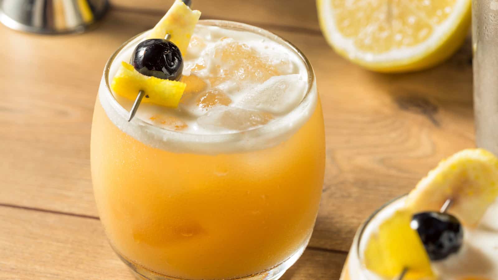 A glass filled with a frothy orange cocktail sits on a wooden surface, garnished with a skewer holding a cherry and pineapple slice. A halved lemon is visible in the background.