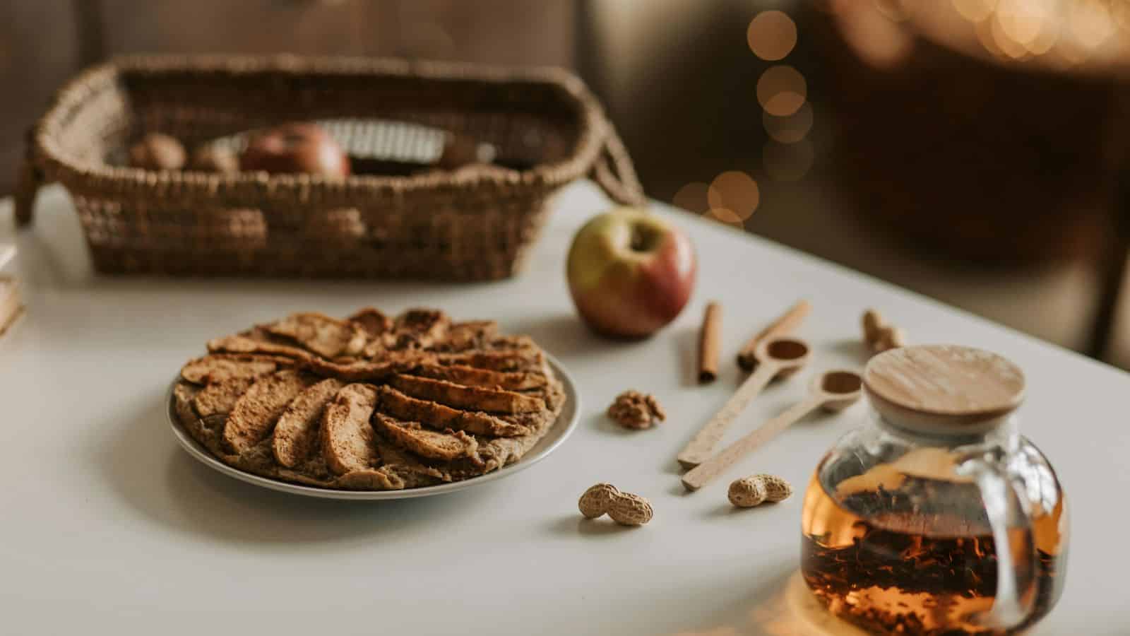 A table with a plate of sliced apple pie, an apple, walnuts, wooden measuring spoons, and a teapot containing tea. There's a woven basket with apples in the background on a couch.