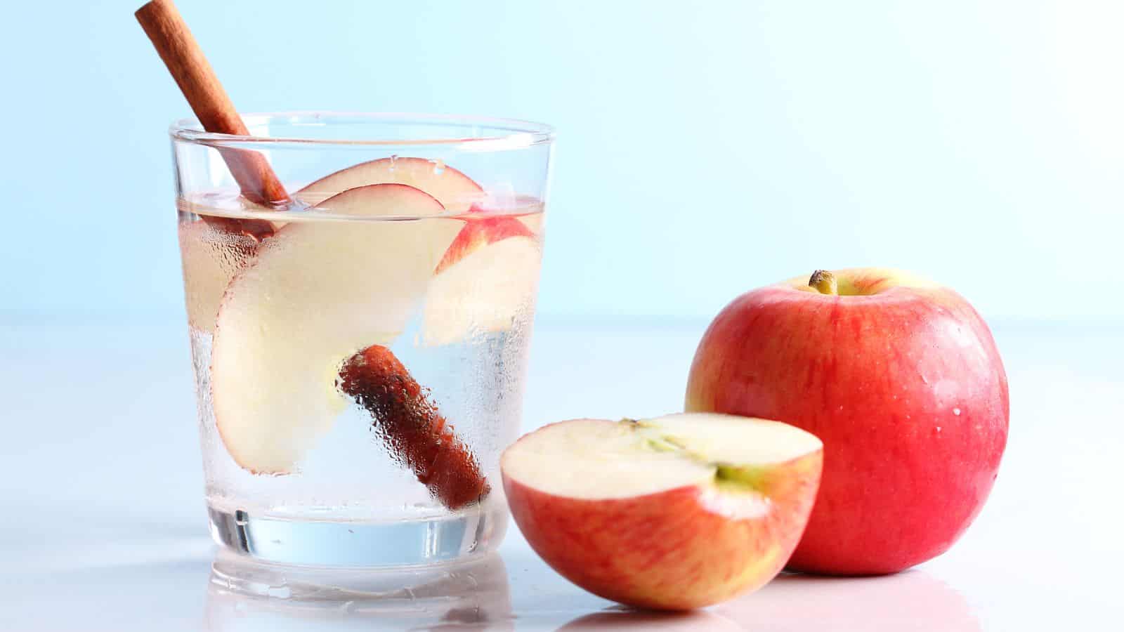 A glass of water with apple slices and a cinnamon stick on a white surface. Next to the glass are two red apples, one whole and one halved. The background is a light blue.