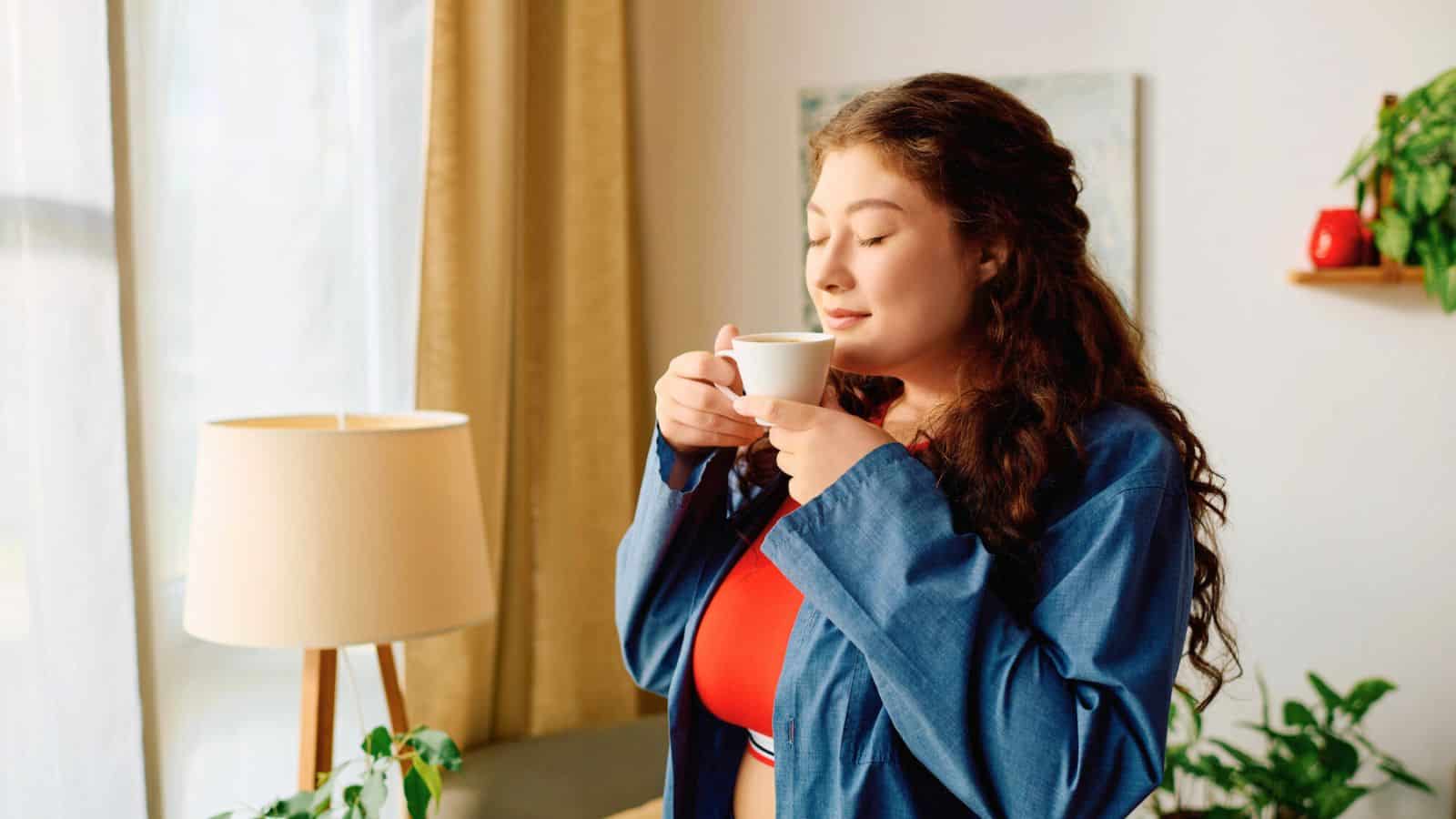 A woman with long brown hair wearing a blue jacket holds a white cup near her face with closed eyes, appearing content. She stands in a well-lit room with plants, a lamp, and a curtain in the background.