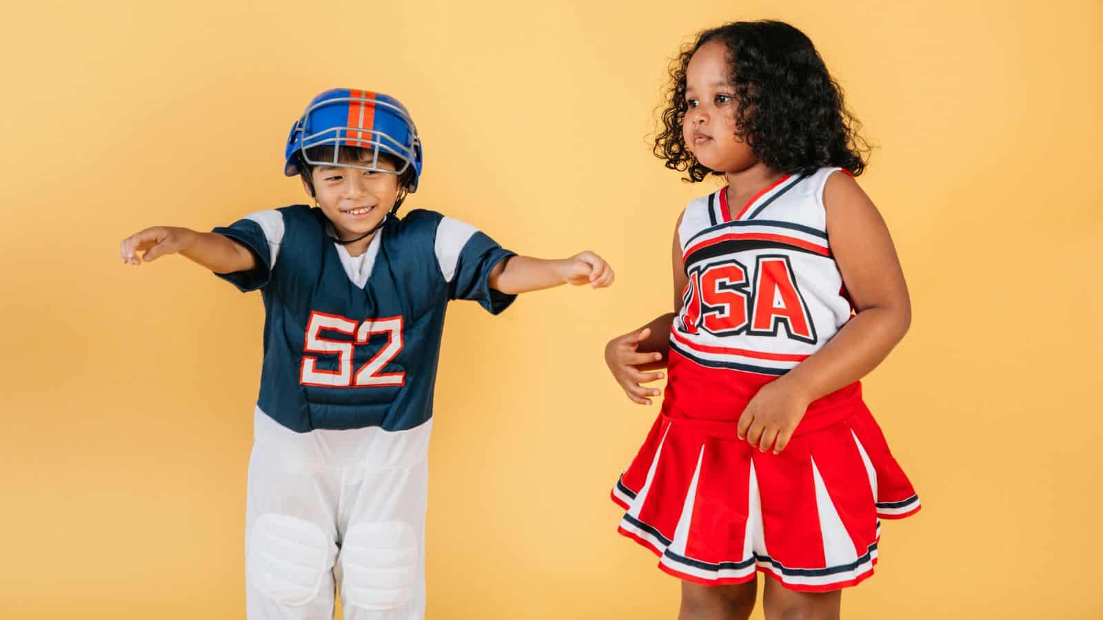 Two children in costumes stand in front of a yellow background. The child on the left wears a football uniform and helmet and has arms outstretched. The child on the right wears a red and white cheerleader outfit labeled "USA.