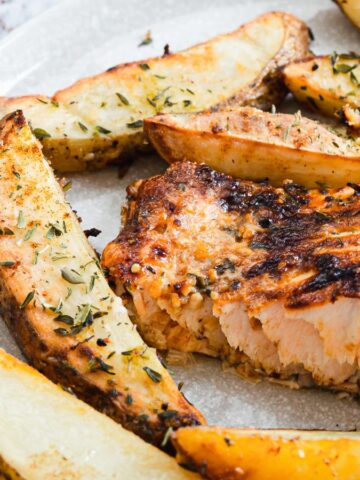 A plate with roasted salmon and herb-seasoned potato wedges. The salmon is flaky with a crispy exterior, and the potato wedges are golden brown. A gray napkin and utensils are partially visible beside the plate.