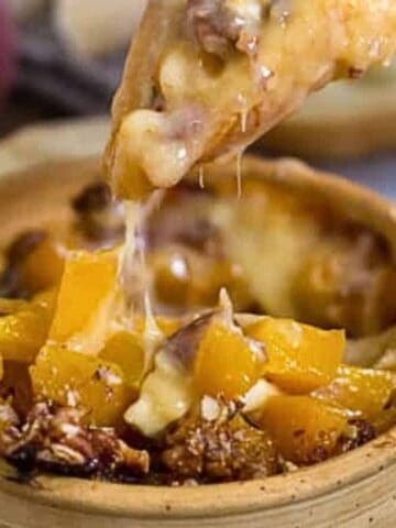 A wooden bowl filled with a cheesy dip featuring diced peaches and pecans. A hand is dipping a piece of bread into the gooey mixture. Slices of bread and pecans are scattered in the background.