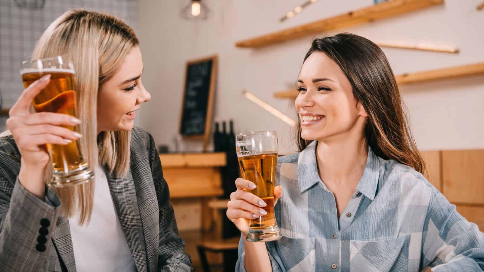 Two women sit at a table in a casual setting, each holding a glass of beer. They are smiling and making eye contact with each other. The background includes wooden decor and a partially visible chalkboard.