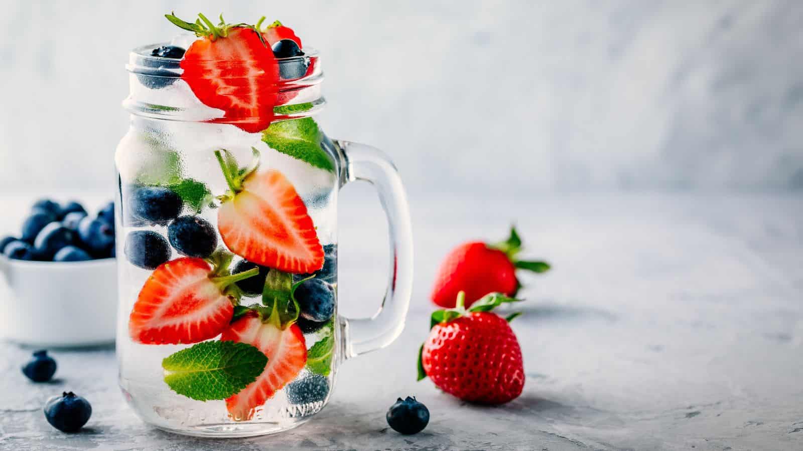 A glass jar filled with water, sliced strawberries, blueberries, and mint leaves sits on a gray surface. A few strawberries and blueberries are scattered outside the jar. A small white bowl with blueberries is in the background.