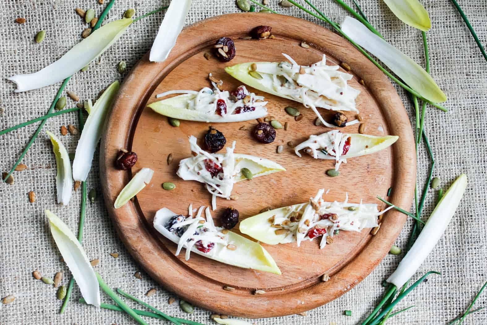 A wooden platter with endive leaves topped with shredded cheese, nuts, and seeds. The platter is placed on a burlap surface, surrounded by scattered endive leaves, nuts, seeds, and chives.