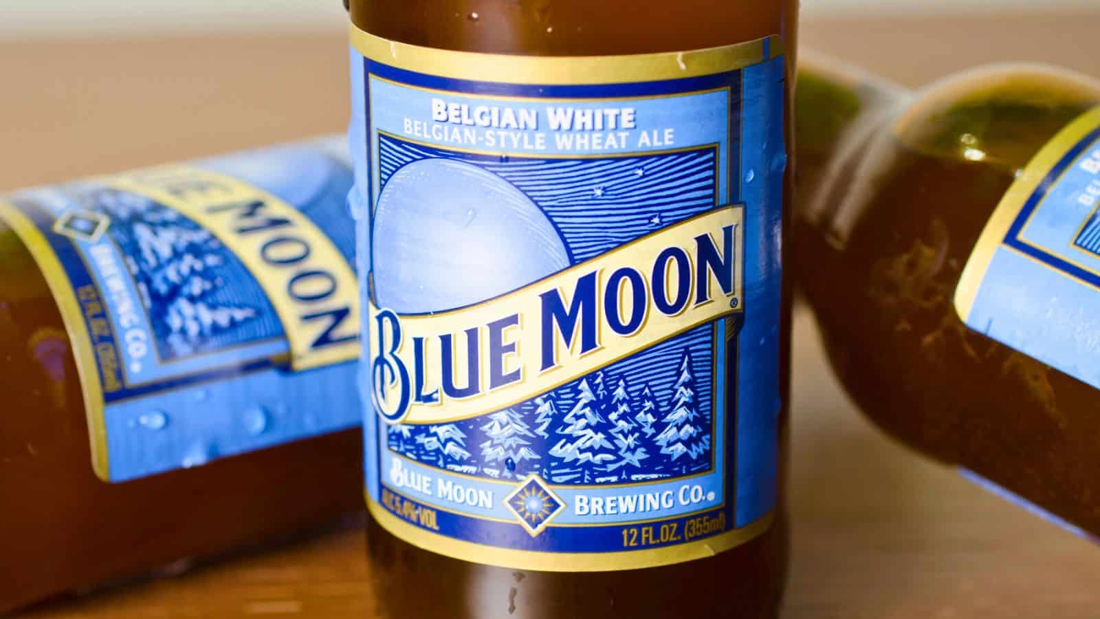 A close-up of a Blue Moon Belgian White beer bottle. The label is blue with white and gold accents, featuring a crescent moon and snow-covered trees. Two additional bottles are partially visible in the background.