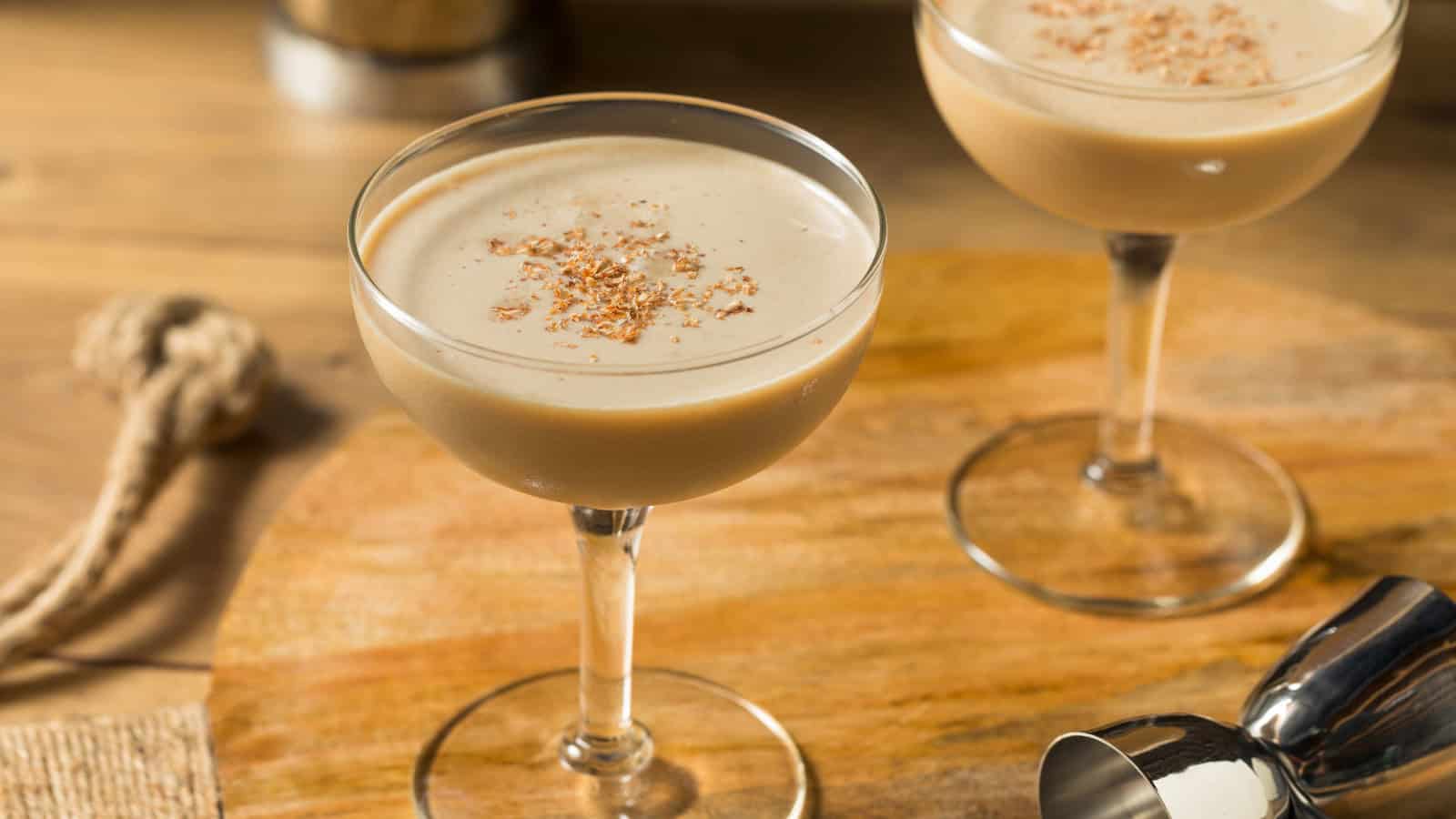 Two coupe glasses filled with a creamy brown cocktail are placed on a wooden surface. Each drink is garnished with a sprinkle of grated spice. A metal cocktail shaker is partially visible in the foreground.