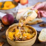 A hand holds a slice of bread dipped in a cheesy Brie dip with peach and pecan mixture from a bowl. Another bowl of peaches and sliced bread are visible in the background. Peaches and pecans are scattered around on a wooden surface, enhancing the rustic ambiance.