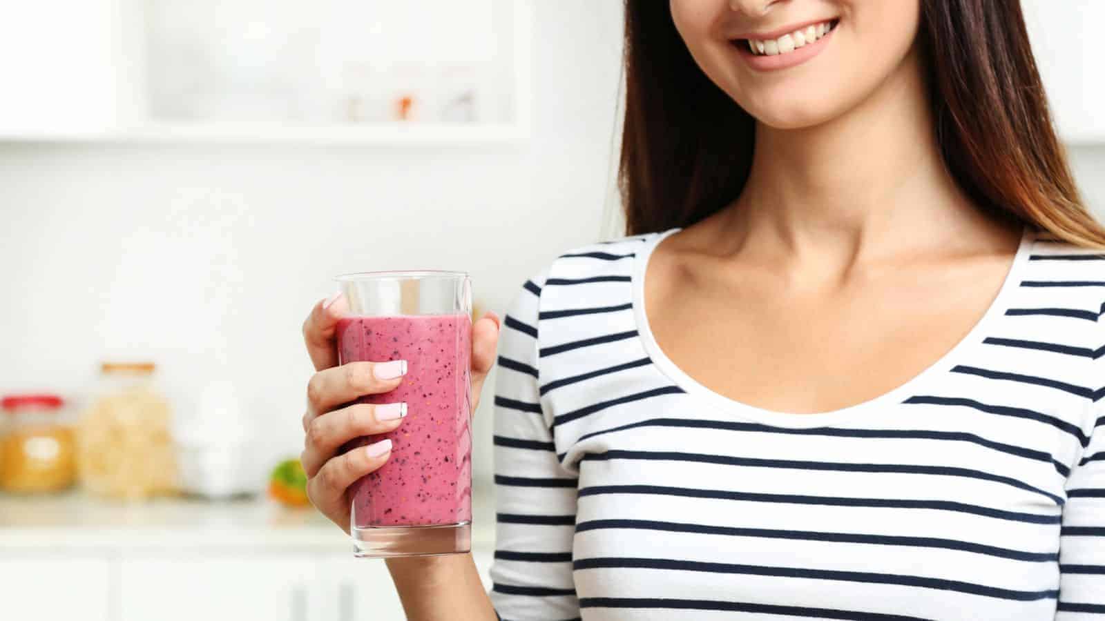 A woman in a black and white striped shirt is smiling and holding a glass of pink smoothie in a kitchen setting. The background is blurred, showing kitchen elements like jars and fruits.