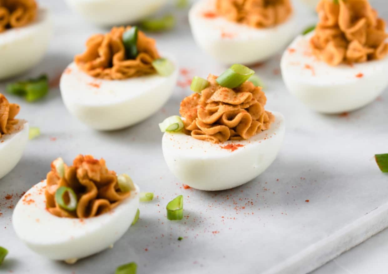 A tray of deviled eggs topped with a creamy orange mixture, garnished with paprika and chopped green onions, arranged neatly on a light-colored surface.