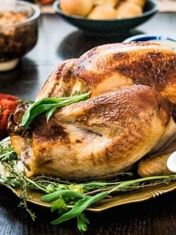 A roasted turkey on a platter is surrounded by side dishes, including a pie, cranberry sauce, green beans, and a gravy boat on a table setup for a meal.